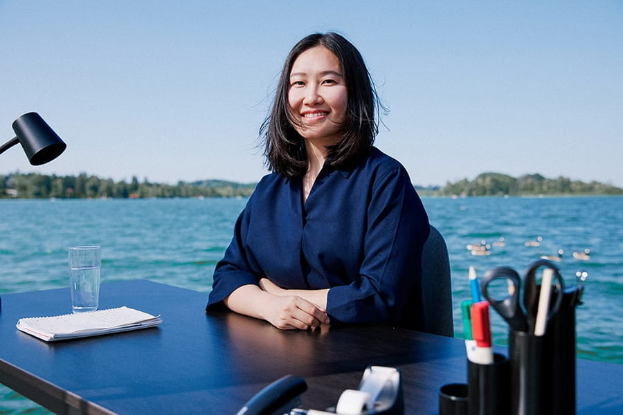 An Asian woman sits at a desk, with a lake in the background.
