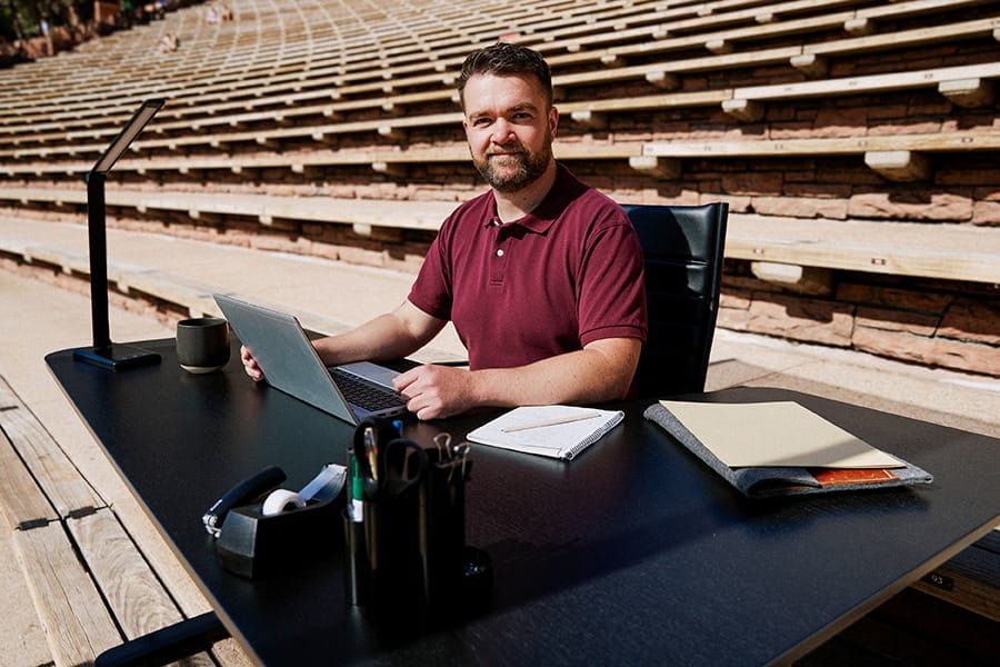 A man sits at a desk, with the stony rows of an amphitheatre in the background.
