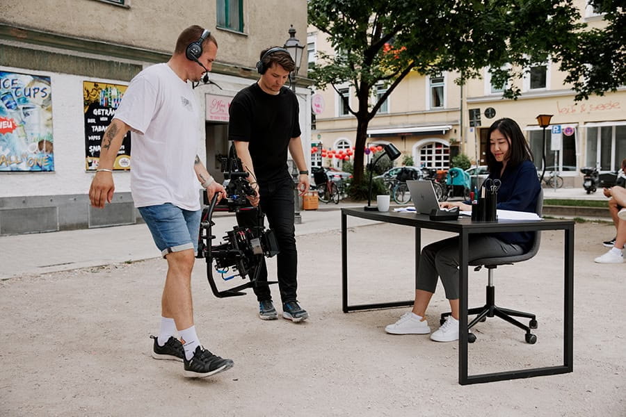 An Asian woman is filmed by two cameramen in an urban environment.