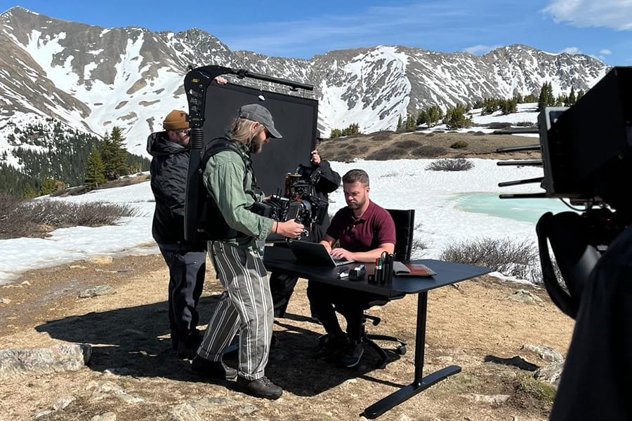 A man is sitting at a desk in a snowy landscape, being filmed by a cameraman.