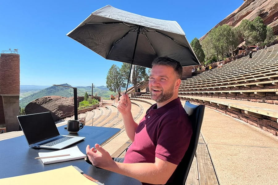 A smiling man sits at a desk in an amphitheatre with an umbrella to protect him from the sun.